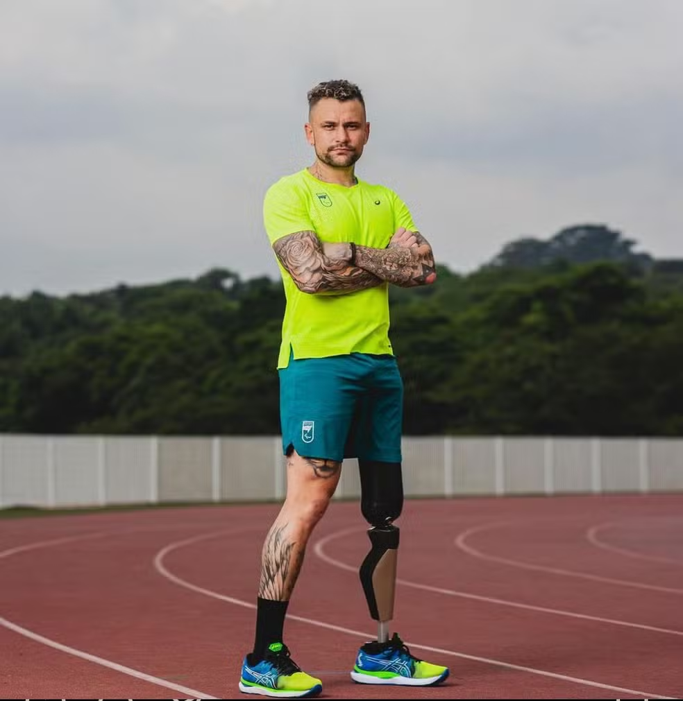 Em uma pista de atletismo, Vinicius Rodrigues está em pé e de braços cruzados. Ele usa uma prótese na perna esquerda.