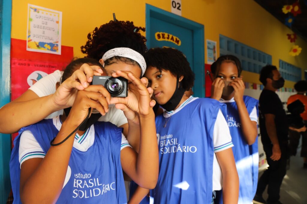 #Pratodosverem Quatro alunos do Instituto Brasil Solidário na Oficina de Fotografia. Um deles segura a máquina na altura dos olhos com ajuda de outro aluno,. Os outros dois observam.