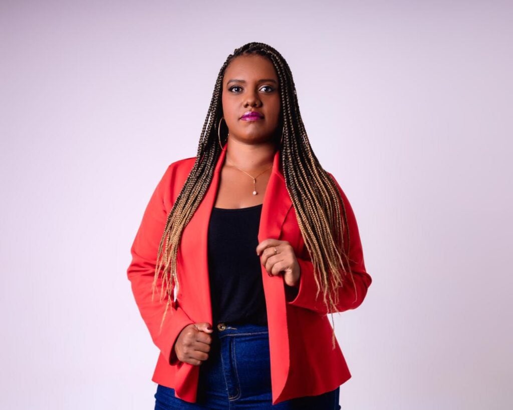 Foto de Flávia Silva. Ela é uma mulher negra de cabelos compridos e aparece da cintura para cima. Usa blazer vermelho sobre camiseta preta e está de calça jeans.