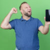 joyful adult slavic man showing mobile phone and doing yes gesture with closed eyes isolated on green background
