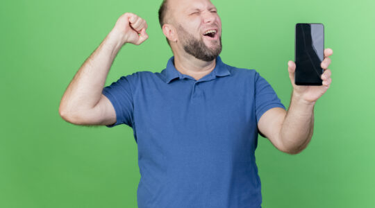 joyful adult slavic man showing mobile phone and doing yes gesture with closed eyes isolated on green background