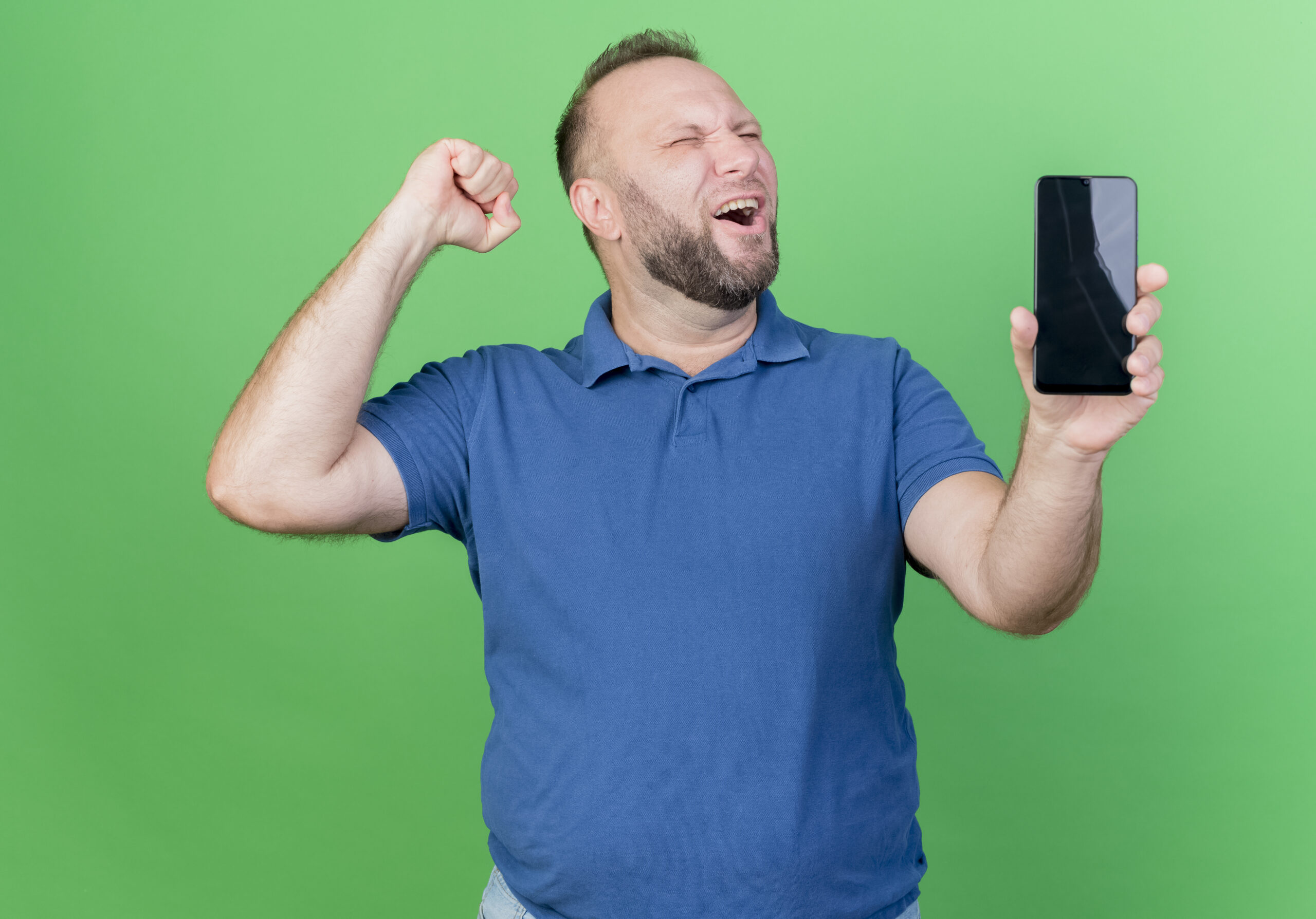 joyful adult slavic man showing mobile phone and doing yes gesture with closed eyes isolated on green background