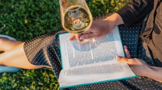Woman with book and wine glass sitting on the grass in the park on the sunset