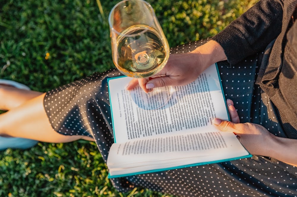 Woman with book and wine glass sitting on the grass in the park on the sunset