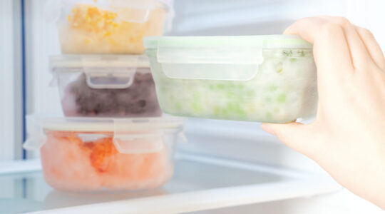Female hand holds a glass container with green peas against the background of freezer with other boxes.
