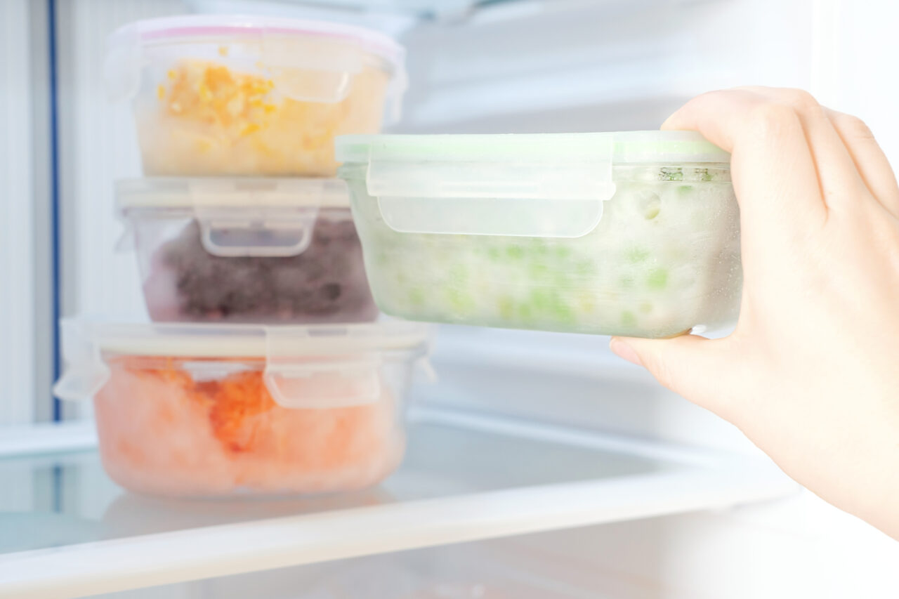 Female hand holds a glass container with green peas against the background of freezer with other boxes.