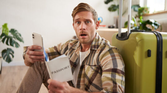 Man with scared face, sits on floor near suitcase, holds smartphone and passport