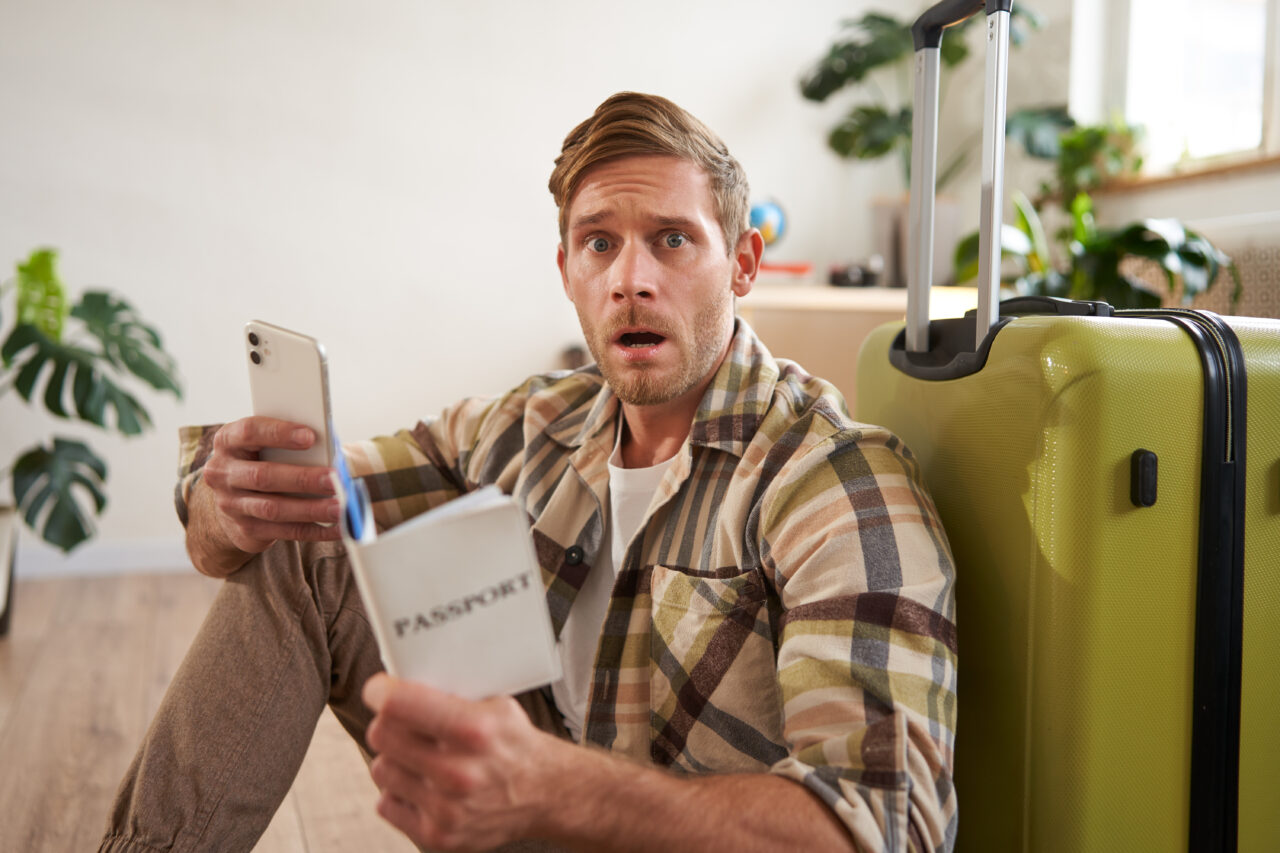 Man with scared face, sits on floor near suitcase, holds smartphone and passport