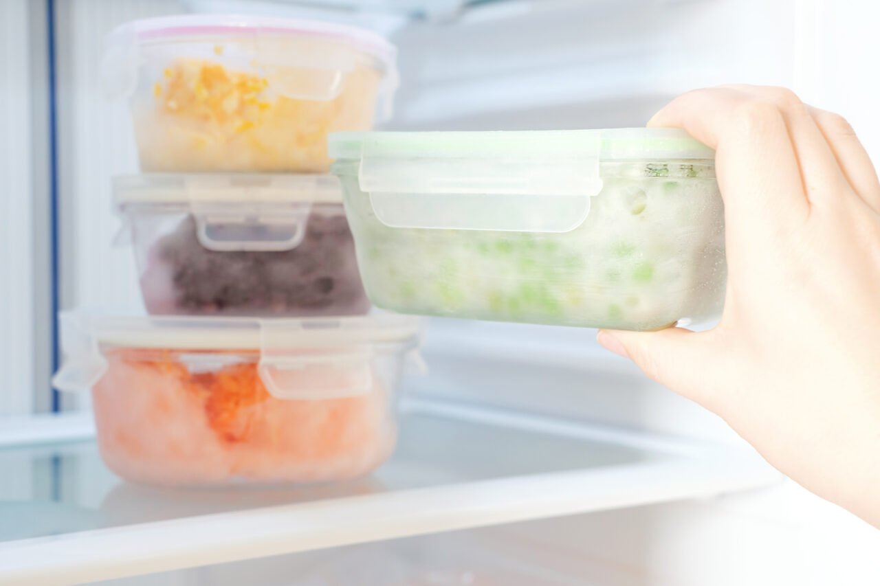 Female hand holds a glass container with green peas against the background of freezer with other boxes.