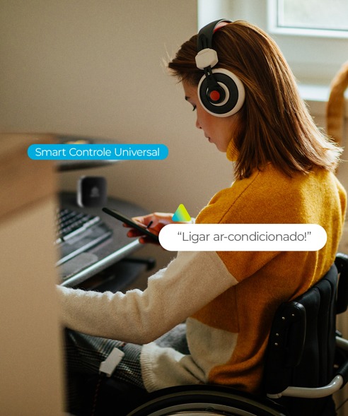 Foto ilustrativa Positivo Casa Inteligente #PraTodosVerem Foto de uma mulher em cadeiras de rodas. De costas para nós, ela está em frente a um notebook sobre uma mesa. Usa fone de ouvido e segura um controle na mão direita, Sobre a foto duas caixas de diálogo: Smart Controle Universal e “Ligue ar-condicionado!”.