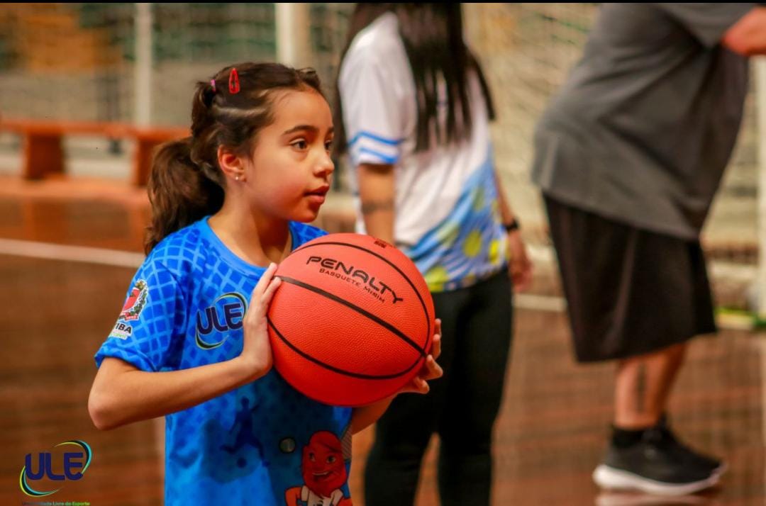 Em um ginásio, menina de cerca de oito anos com a camiseta da ULE na cor azul. A menina segura uma bola de basquete.