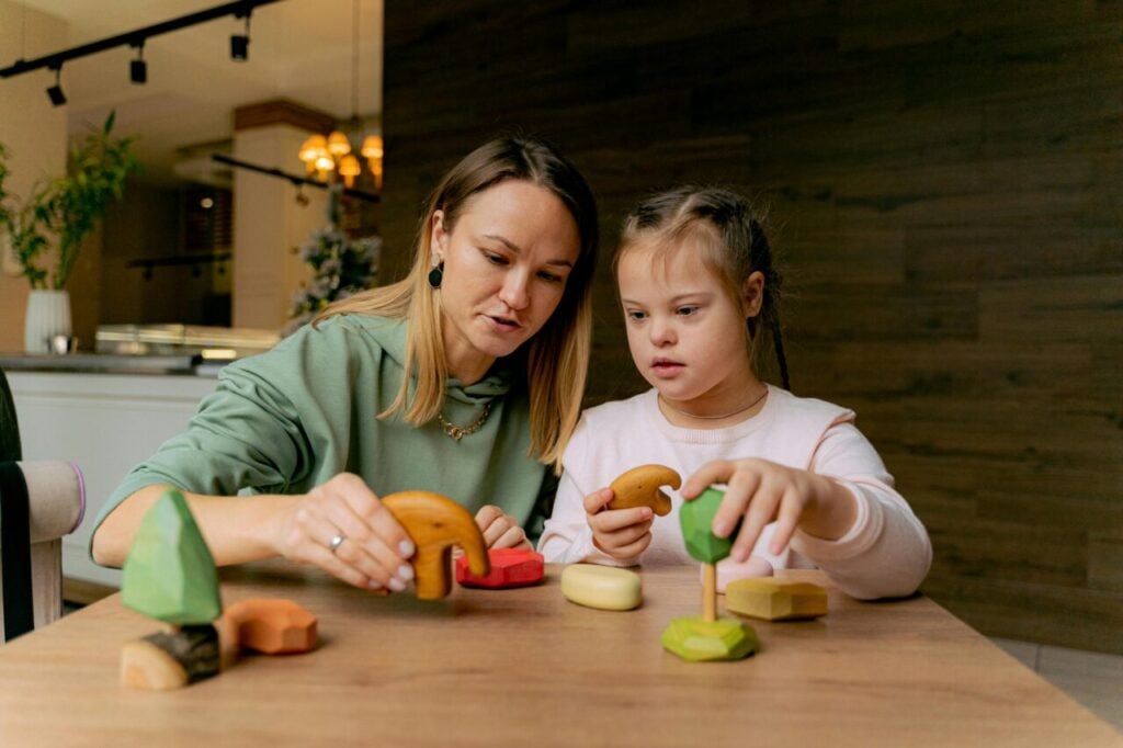 Mulher e criança brancas, em frente a uma mesa com dezenas de objetos em madeira. A criança, com características da Síndrome de Down, olhos puxados e rosto arredondado, olha atentamente para um objeto no formato de um elefante, nas mãos da mulher.