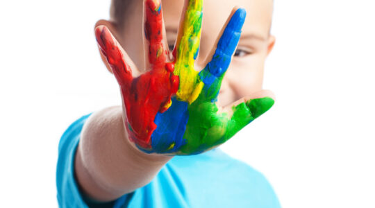 Criança com camiseta azul espalma, em frente ao rosto, mão direita com tinta nas cores vermelho, azul, amarelo e verde.