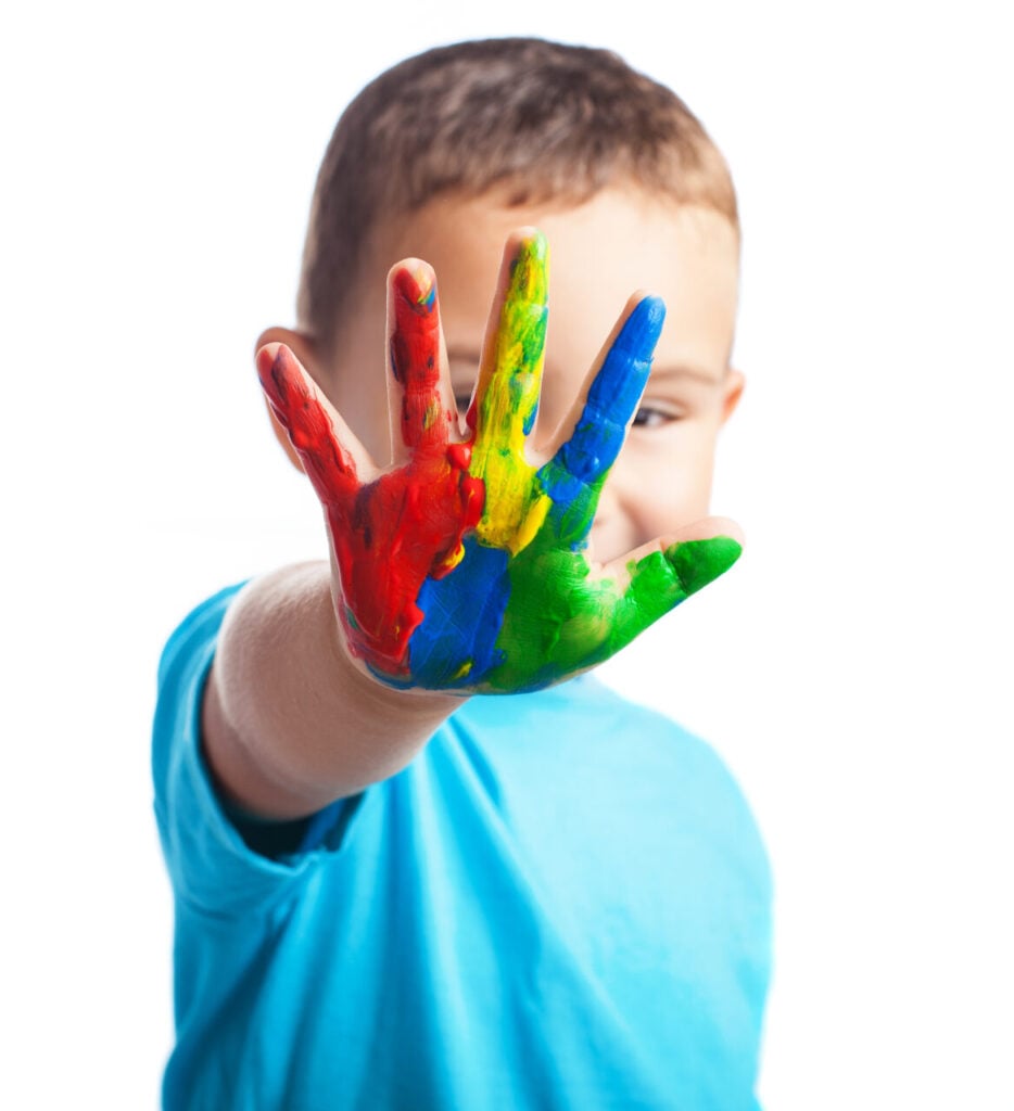 Criança com camiseta azul espalma, em frente ao rosto, mão direita com tinta nas cores vermelho, azul, amarelo e verde.