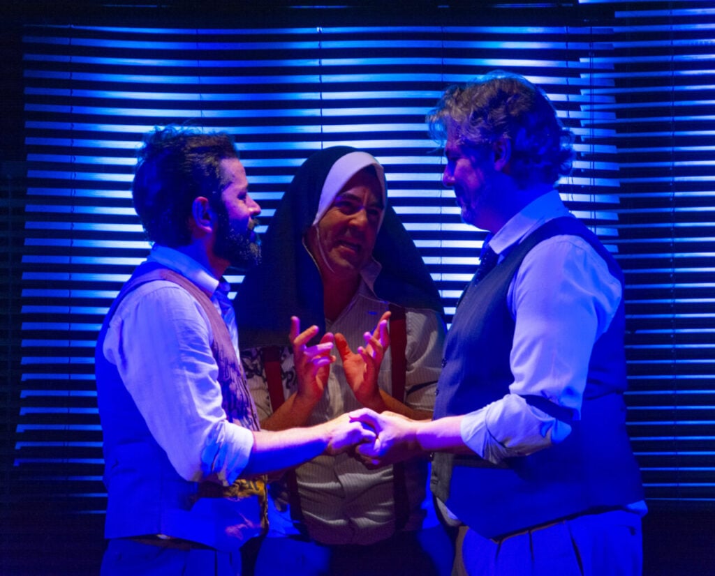 Iluminados de azul, em frente a uma persiana, 3 pessoas. Um casal de homens frente à frente de mãos dadas. No centro, entre eles, uma freira.