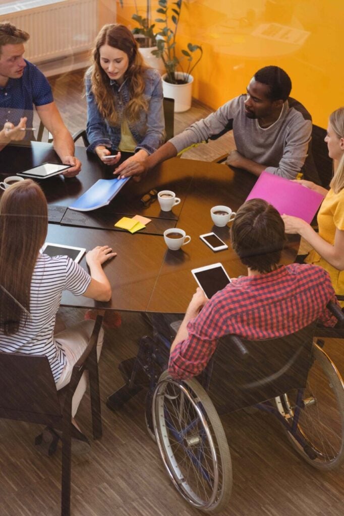 Em uma sala, envolta de uma mesa redonda, seis jovens de diversas etnias, entre eles, de costas para nós, uma pessoa em cadeira de rodas.