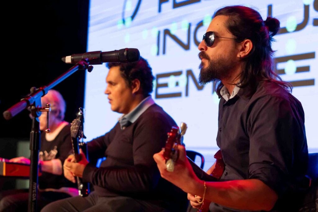 Foto horizontal de Arthur Guerino. Em um palco, três músicos tocam sentados. O primeiro toca teclado, o segundo e o terceiro guitarra. Ao fundo, um telão branco com letras pretas.