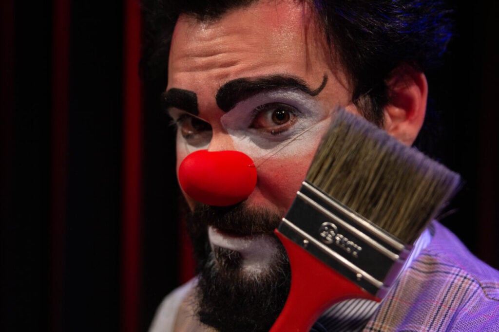 Foto do rosto de Olirrô, o Pintor. Ele é um homem branco de cabelos, barba e bigodes pretos. Tem as sobrancelhas grossas e delineadas e nariz de palhaço. O rosto é pintado de branco na região dos olhos e da boca. Ele mostra à frende dele, um pincel.