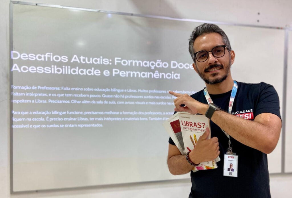 Em uma sala, ao lado de um telão de fundo preto e letras brancas, um homem branco e sorridente de camiseta preta e com três livros na mão. Ele aponta para o telão com frase, em branco: Desafios Atuais: Formação Docente Acessibilidade e Permanência.