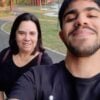 Na foto vertical, Mateus em primeiro plano, ligeiramente inclinado para frente. Ele tem barba curta e cabelos pretos. Usa veste uma camiseta preta e sorri. Atrás dele e à esquerda, está Viviane, uma mulher de cabelos castanhos escuros e lisos, na altura dos ombros. Usa uma blusa preta e uma alça de bolsa vinho atravessa seu ombro. Ela também sorri. Ao fundo, há uma área de parque, com árvores altas, gramado e uma pista asfaltada.