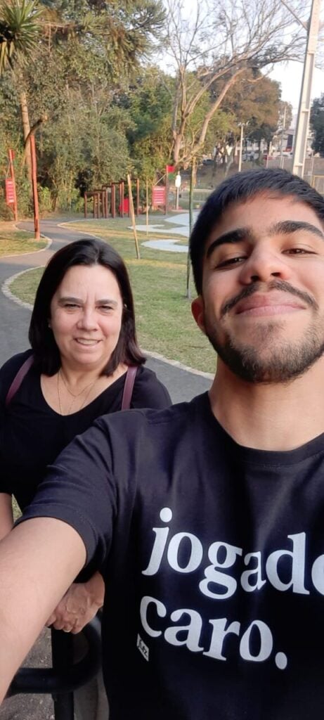 Na foto vertical, Mateus em primeiro plano, ligeiramente inclinado para frente. Ele tem barba curta e cabelos pretos. Usa veste uma camiseta preta e sorri. Atrás dele e à esquerda, está Viviane, uma mulher de cabelos castanhos escuros e lisos, na altura dos ombros. Usa uma blusa preta e uma alça de bolsa vinho atravessa seu ombro. Ela também sorri. Ao fundo, há uma área de parque, com árvores altas, gramado e uma pista asfaltada.