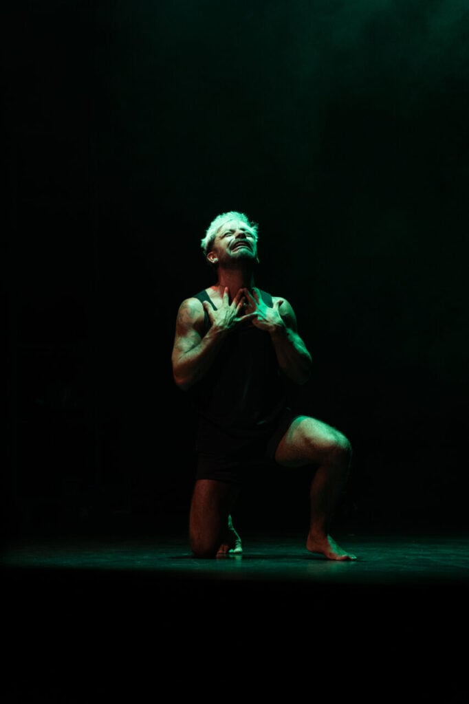 Em um palco escuro de chão preto, um homem branco de cabelos loiros e barba e bigodes ralos e pretos. Ele usa regata e shorts pretos. Com os olhos espremidos, a cabeça levemente para trás, ele esboça uma expressão de sofrimento com a boca fechada. Está com as mãos espalmadas junto ao peito e com um joelho no chão e o outro flexionado.
