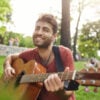 Joyfully singing and playing his favourite songs street musician. Cheerful and smiling bearded guy holding guitar in his hands, touching guitar strings.