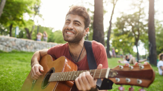 Joyfully singing and playing his favourite songs street musician. Cheerful and smiling bearded guy holding guitar in his hands, touching guitar strings.