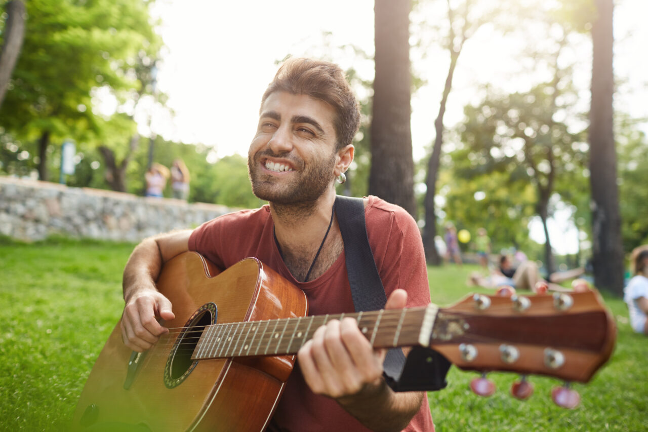 Joyfully singing and playing his favourite songs street musician. Cheerful and smiling bearded guy holding guitar in his hands, touching guitar strings.