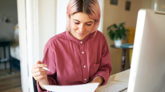 Creative confident young woman sitting in front of computer monitor with papers in her hands, filling in application form, searching for new job. Pretty student girl doing research for course paper