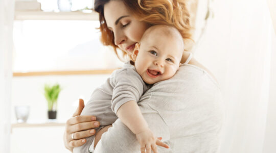 Happy young mom holds precious little child and gently hugging his little body. Kid laughing joyfully and looking in camera with big grey eyes.