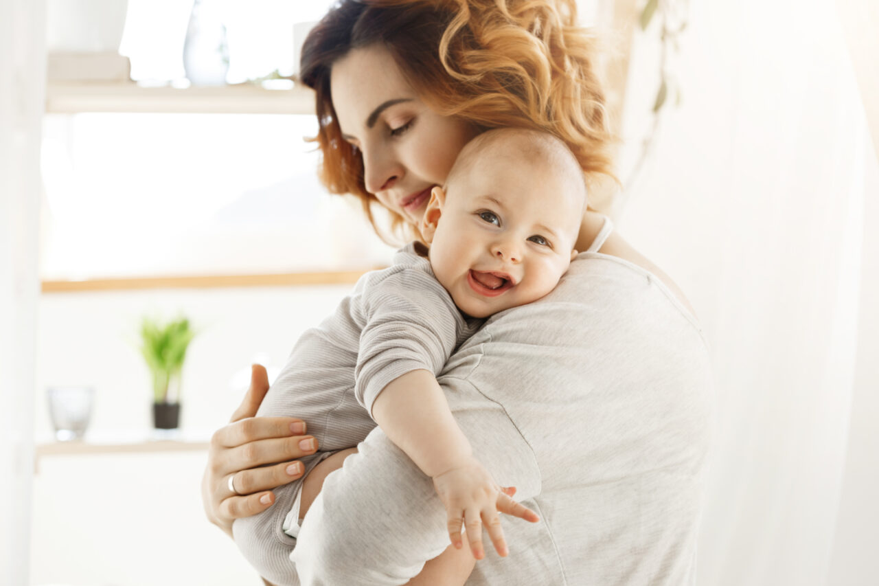 Happy young mom holds precious little child and gently hugging his little body. Kid laughing joyfully and looking in camera with big grey eyes.