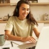 Beautiful young woman in casual clothes using wireless headphones, listening to webinar online on portable computer, sitting in kitchen, drinking tea, writing down. Technology, business and education