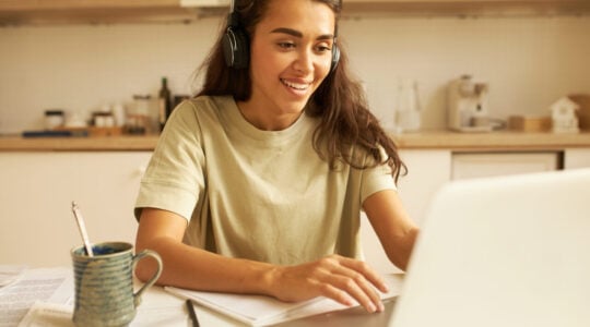 Beautiful young woman in casual clothes using wireless headphones, listening to webinar online on portable computer, sitting in kitchen, drinking tea, writing down. Technology, business and education
