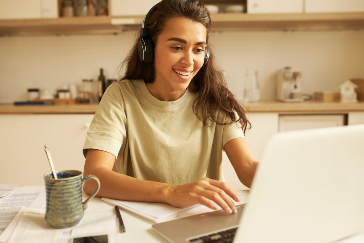 Beautiful young woman in casual clothes using wireless headphones, listening to webinar online on portable computer, sitting in kitchen, drinking tea, writing down. Technology, business and education