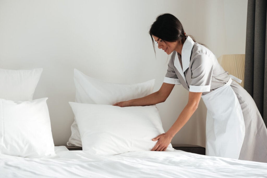 Young hotel maid setting up pillow on bed