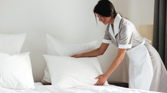 Young hotel maid setting up pillow on bed