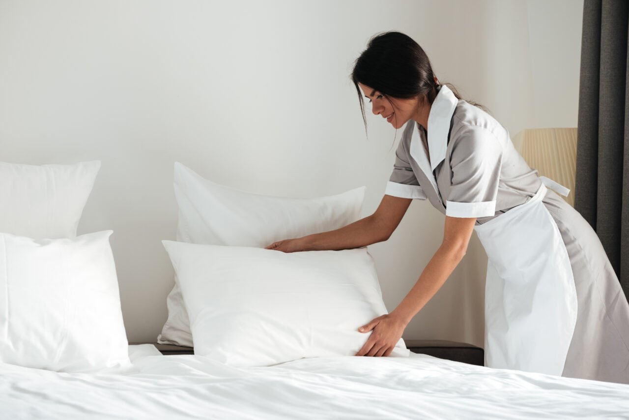 Young hotel maid setting up pillow on bed