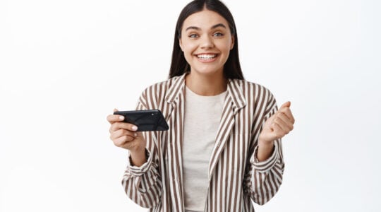 Cheerful young woman winning money on phone, achieve goal in smartphone app, holding mobile phone horizontally and say yes, white background