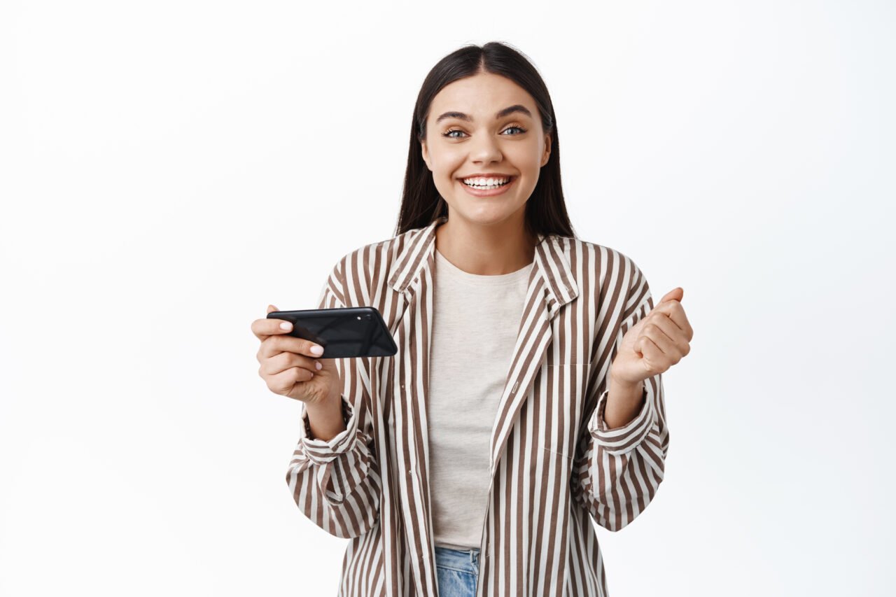 Cheerful young woman winning money on phone, achieve goal in smartphone app, holding mobile phone horizontally and say yes, white background