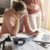 Young housewife dressed casually focused on paperwork, reading document in her hands with concentrated look, managing domestic finances alone while her husband drinking coffee in background