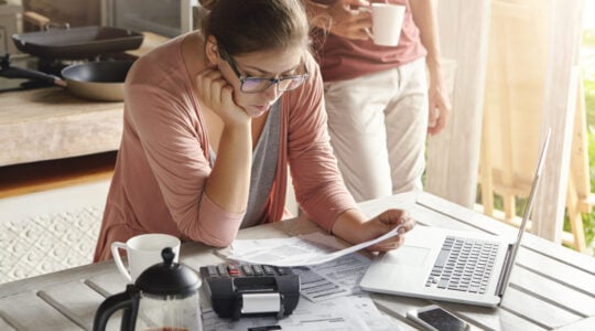 Young housewife dressed casually focused on paperwork, reading document in her hands with concentrated look, managing domestic finances alone while her husband drinking coffee in background