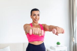 Domestic Training With Weights. Positive black lady doing exercises with dumbbells, strengthening her body at home. Smiling young female working on her biceps muscles, staying healthy