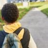 Back view of black child with curly hair carrying backback on shoulders going home from school or sport center on pedestrian road on sunny spring day. Children and elementary education