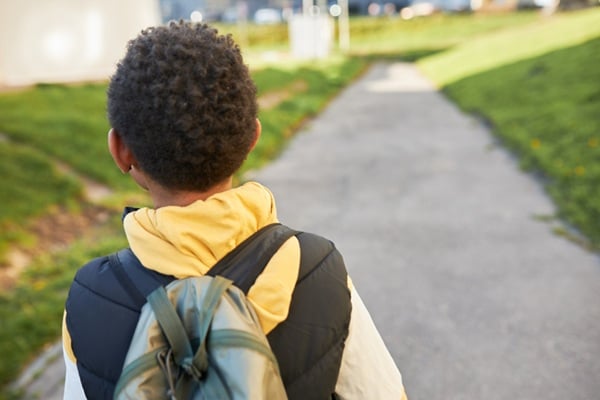 Back view of black child with curly hair carrying backback on shoulders going home from school or sport center on pedestrian road on sunny spring day. Children and elementary education