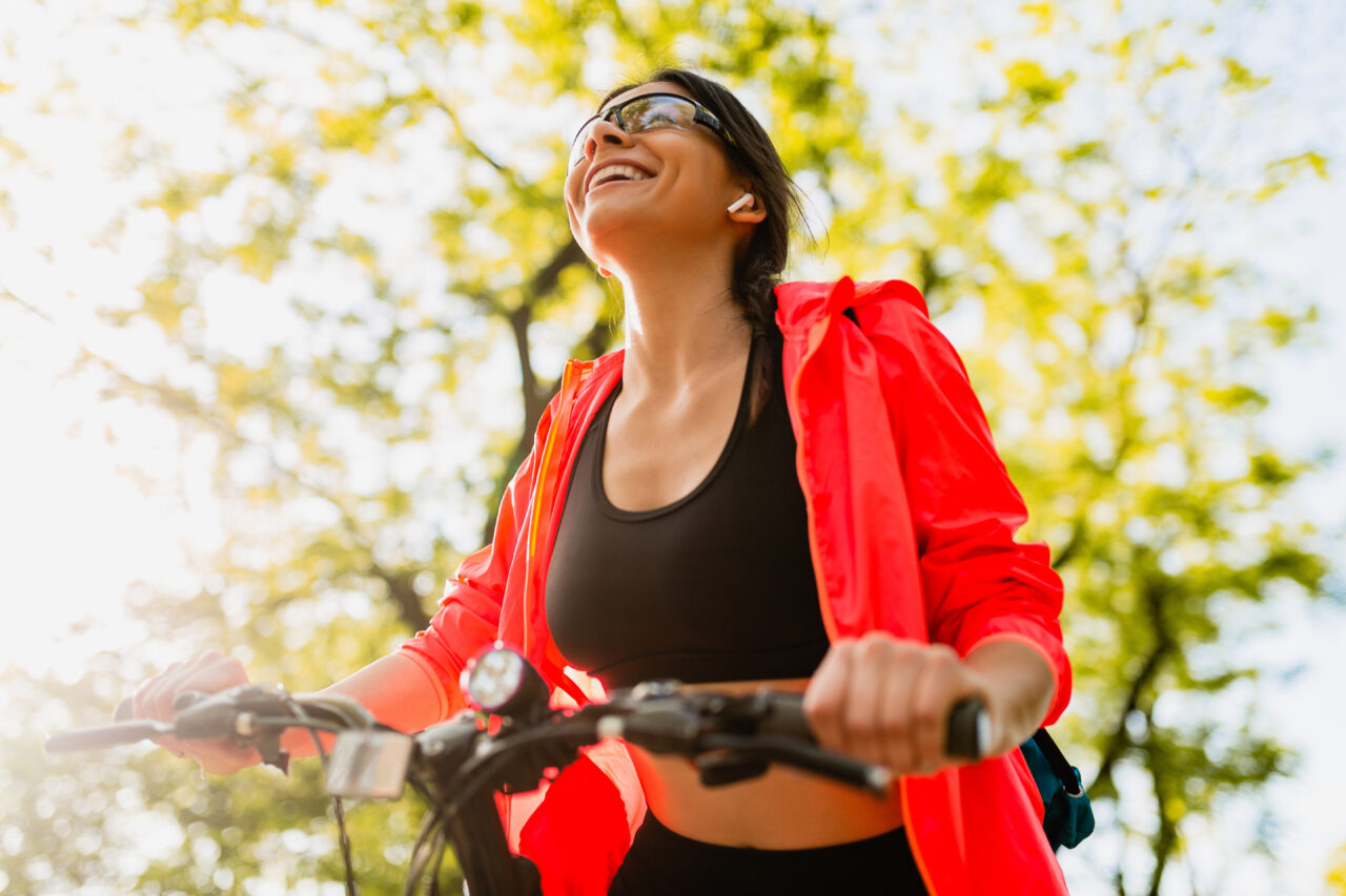 slim fit beautiful woman doing sports in morning in park riding on bicycle on road with yoga mat in colorful fitness outfit, exploring nature, smiling happy healthy lifestyle