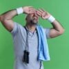 mature sporty man in headband with towel around his neck covering eyes with hands with annoyed expression standing over green background