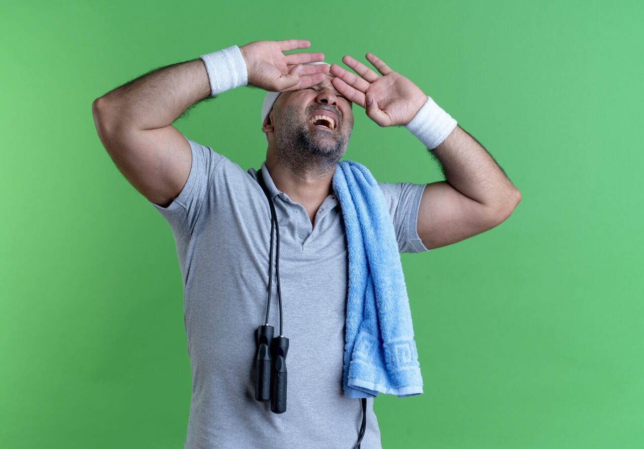 mature sporty man in headband with towel around his neck covering eyes with hands with annoyed expression standing over green background