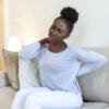 Young Black woman suffering from backache at home. Portrait of a young girl sitting on the couch at home with a headache and back pain. Beautiful woman Having Spinal Or Kidney Pain