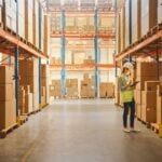 Retail Warehouse full of Shelves with Goods in Cardboard Boxes, Female Worker Checks and Sorts Packages for Delivery. Distribution Logistics Center.
